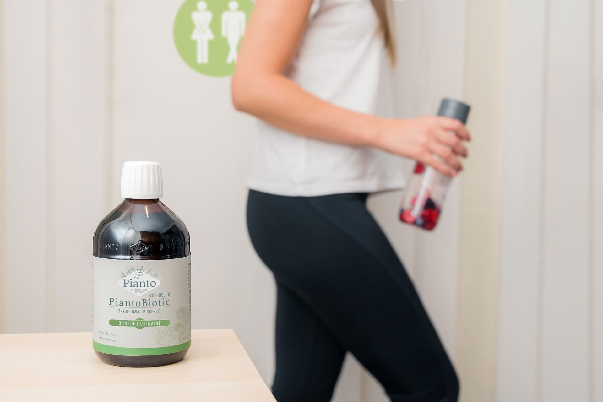 PiantoBiotic Confort Urinaire Bouteille de PiantoBiotic Confort Urinaire posée sur une table. Dans le fond se trouve une jeune fille avec une tenue de sport et une bouteille d'eau à la main