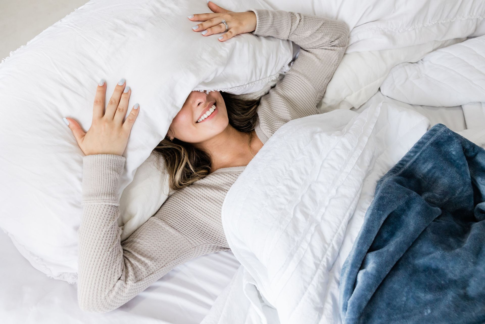 Jeune souriante dans son lit qui tient son oreiller sur la tête 