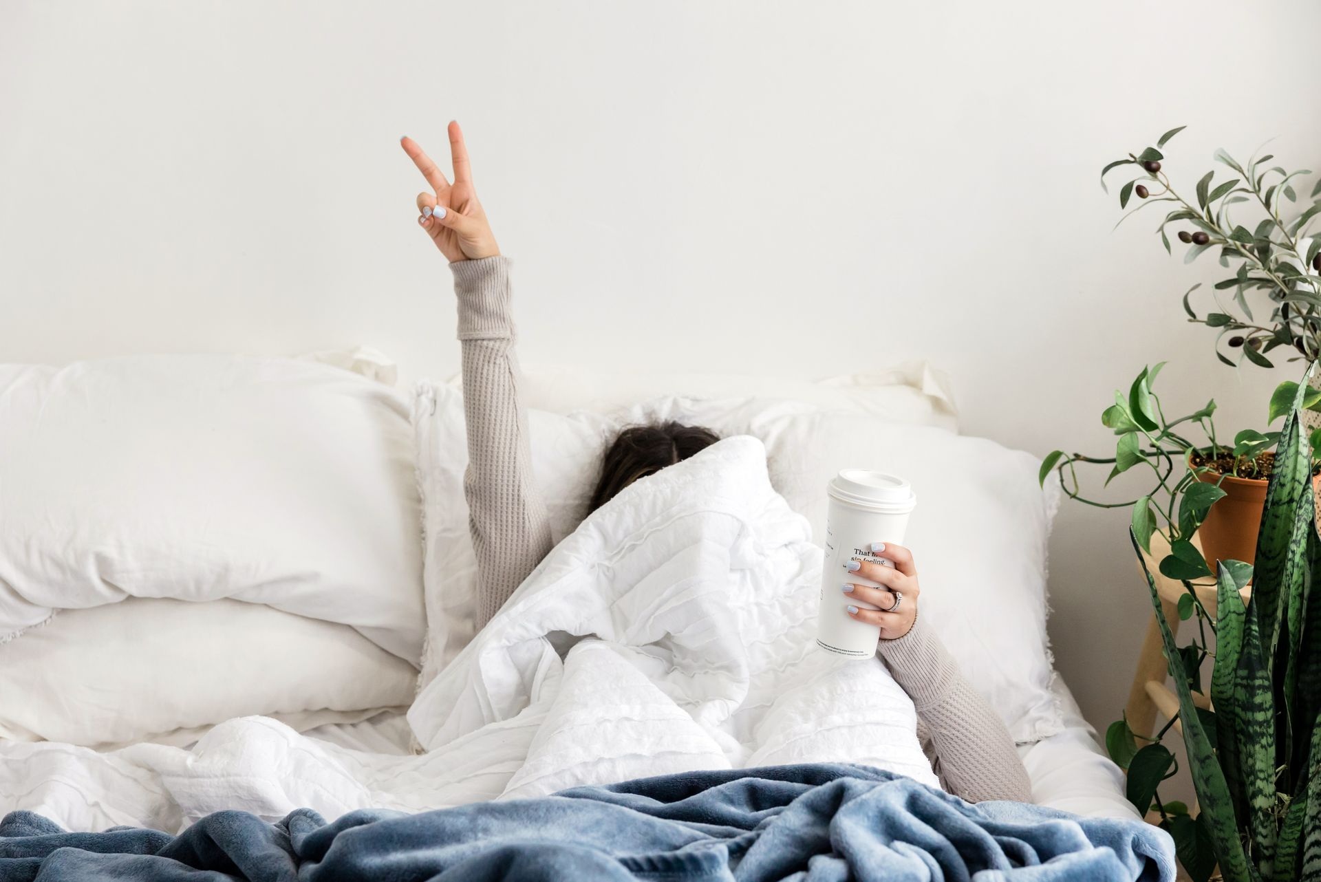 Jeune femme dans son lit avec les doigts en V  et une boisson dans l'autre mainqui se cache derrière sa couette