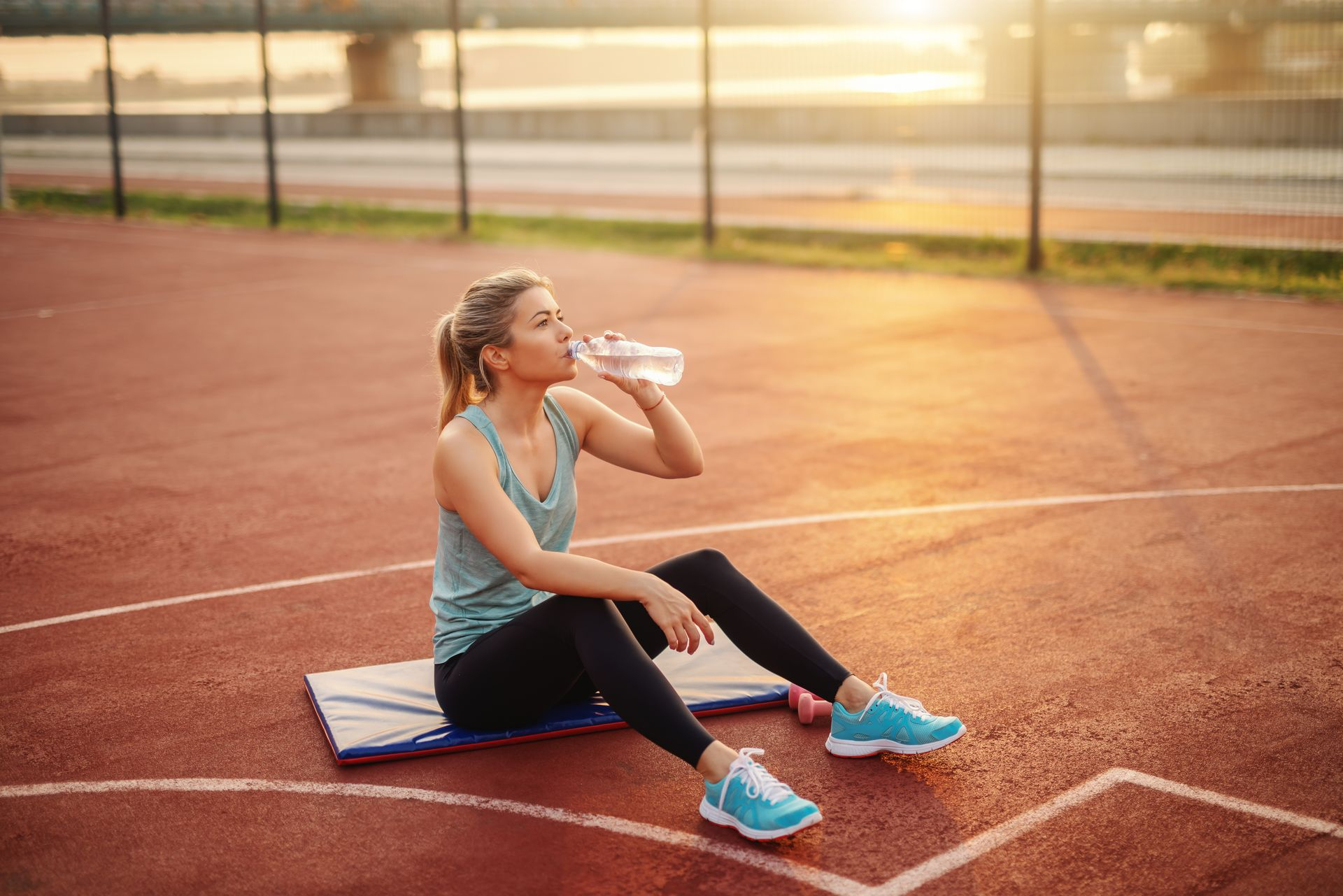Jeune femme en tenue de sport, assise sur un tapis dans un terrain de sport occupée à boire dans une bouteille d'eau 
