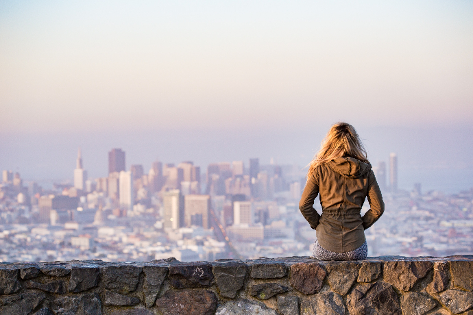 Jeune femme blonde, de dos,  les mains dans les poches, assise sur un mur, occupée à regarder une ville 